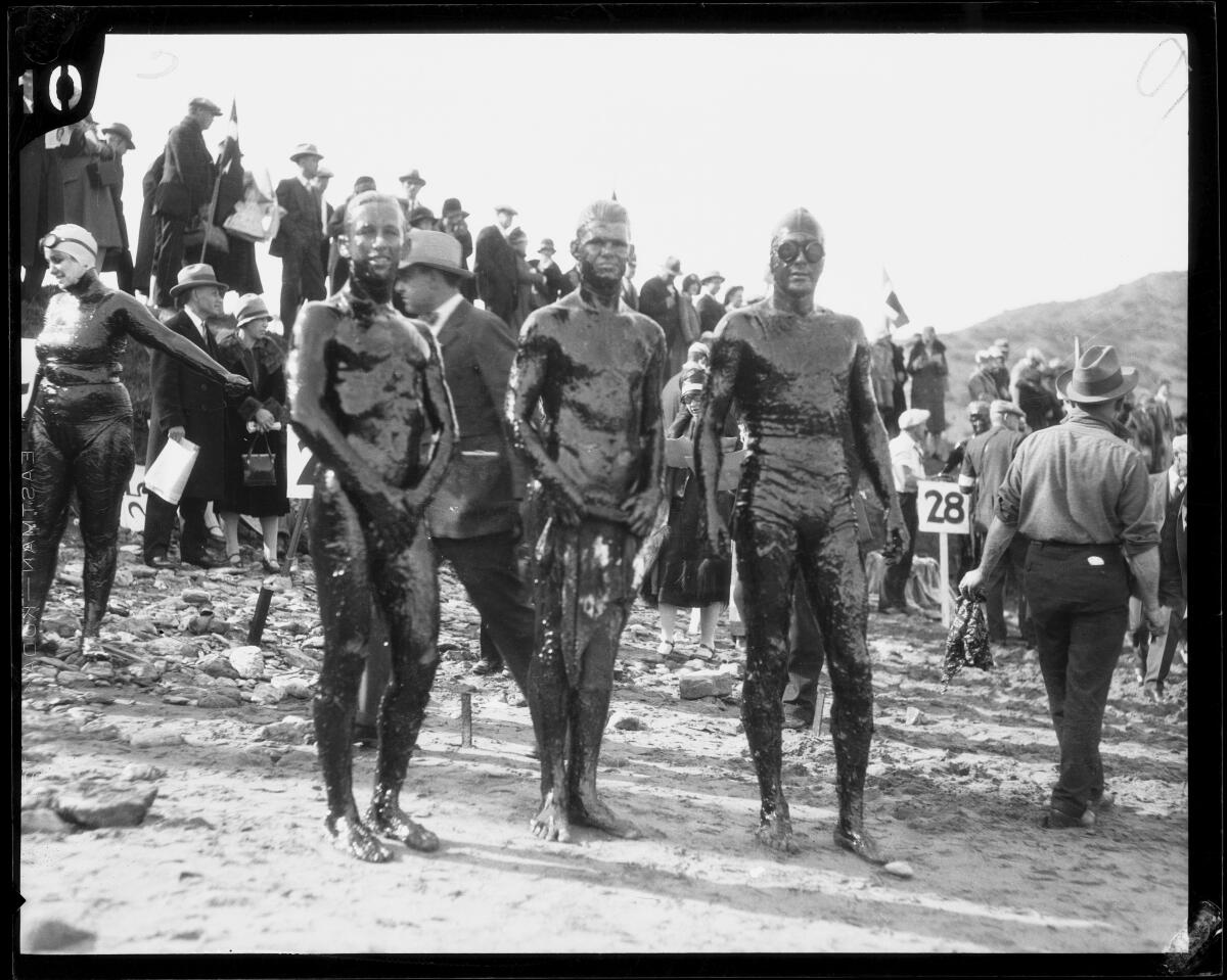 Three people in swim trunks stand on the beach,  covered in oil 