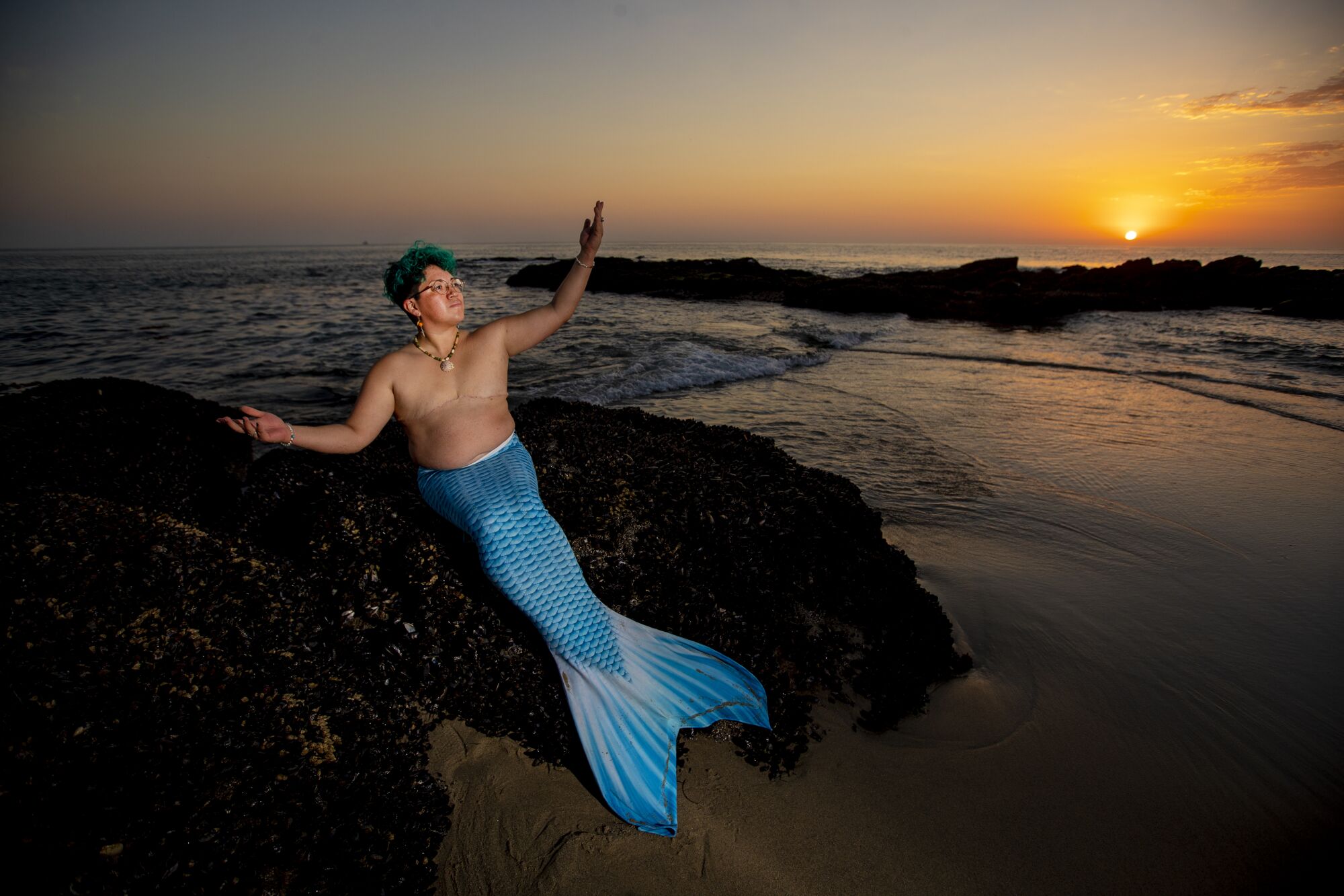 Sammy Silva, wearing full mermaid gear with a tail and no shirt, poses by the beach.