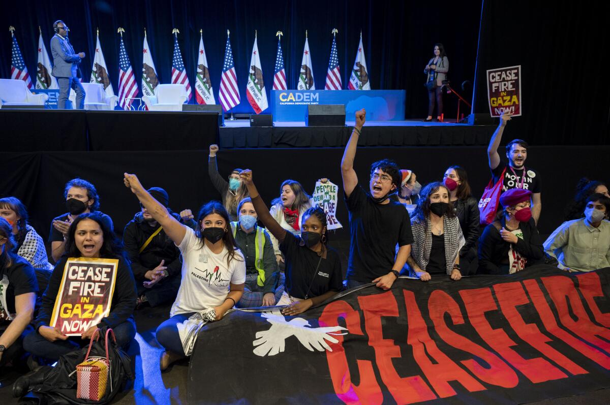 Pro-Palestinian demonstrators sit in front of a stage, some holding a banner that says "ceasefire"