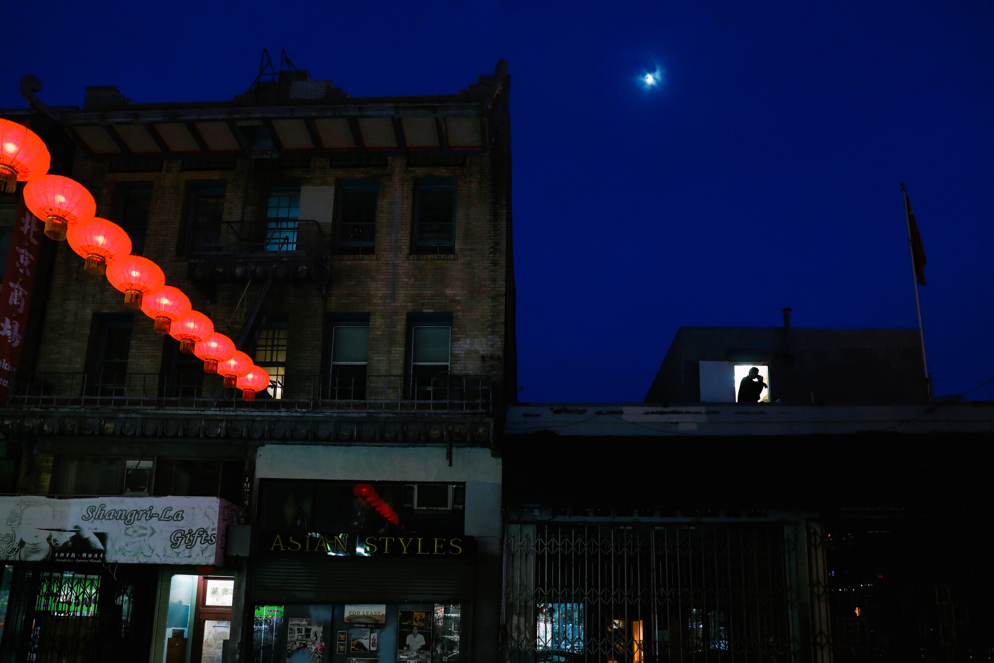Red lanterns glow above a street; higher up, a dot of moon shines.