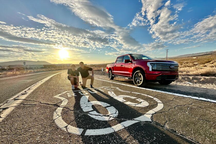 Ford CEO Jim Farley and son kneel on pavement. Red pickup on right.  Route 66 sign painted in white on  highway.