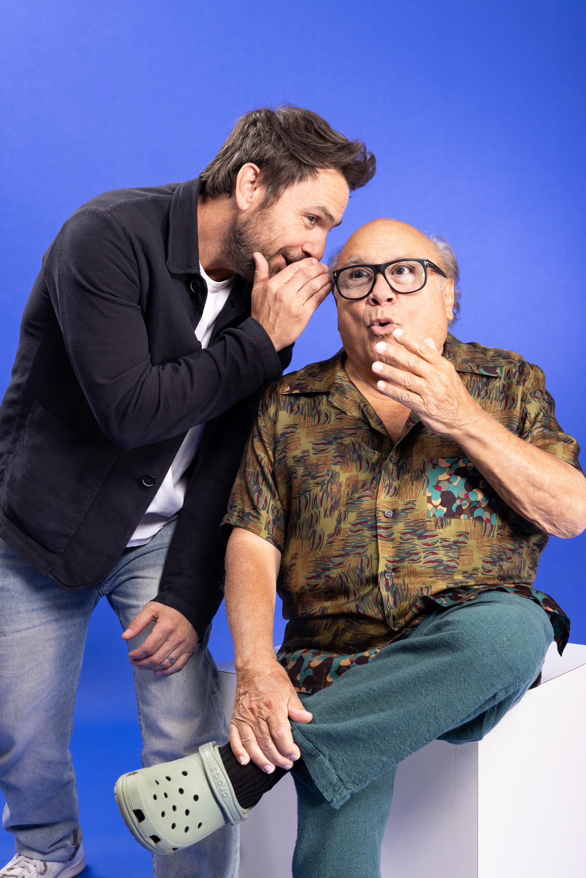 A man in a black jacket and jeans whispers into the ear of an older bald man in glasses sitting on a white cube.