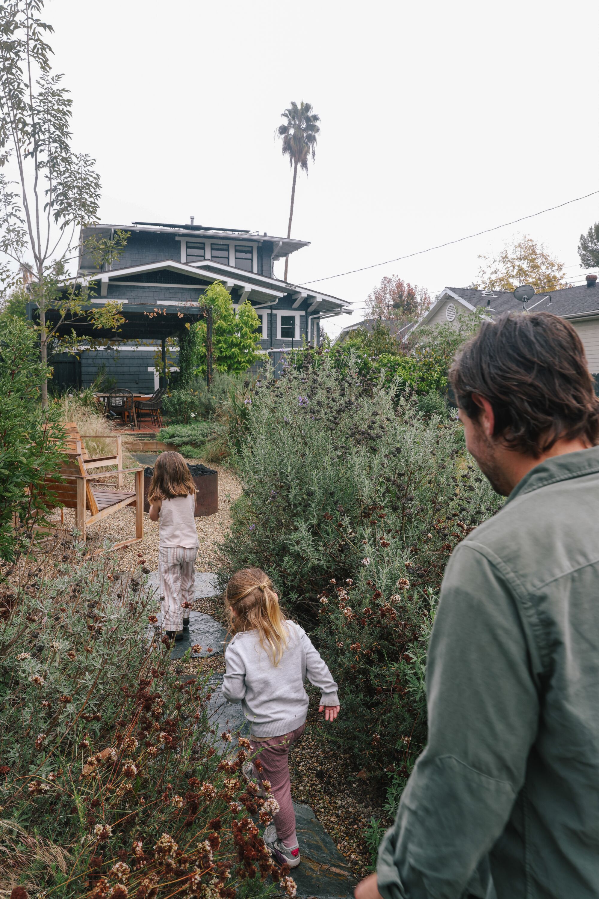 A father and his two young daughters walk through a garden toward a Craftsman house.