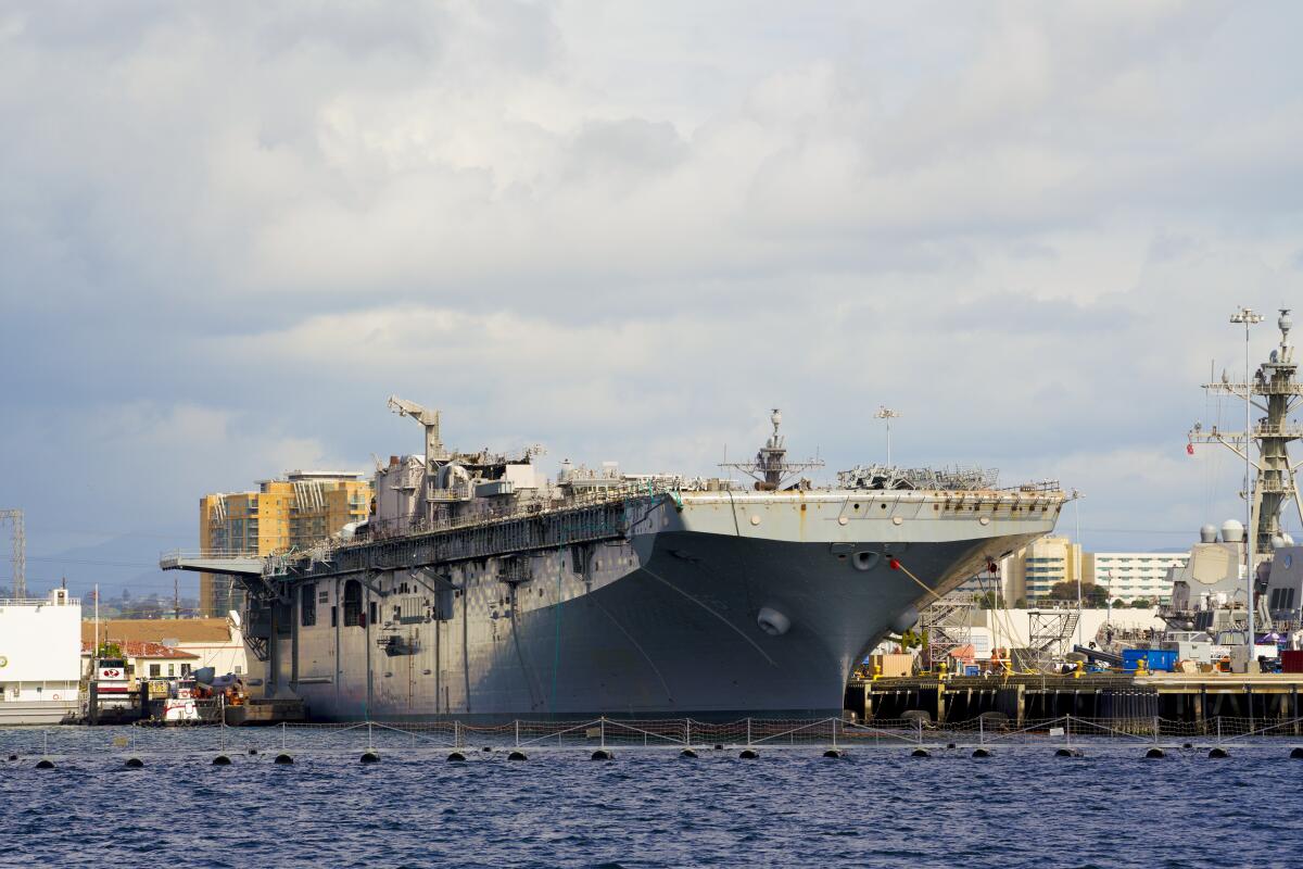 A large Navy ship docked at a pier, its central island structure partially dismantled
