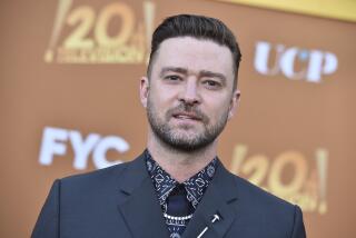 Justin Timberlake sports a short beard, a printed shirt and a black blazer as he poses in front of a promotional backdrop