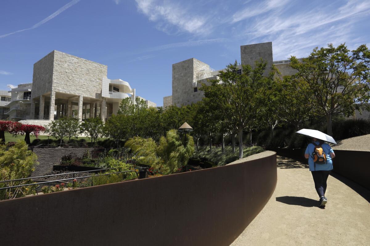 A woman carries an umbrella for shade and walks in the Getty Center garden, with the museum building in the background.