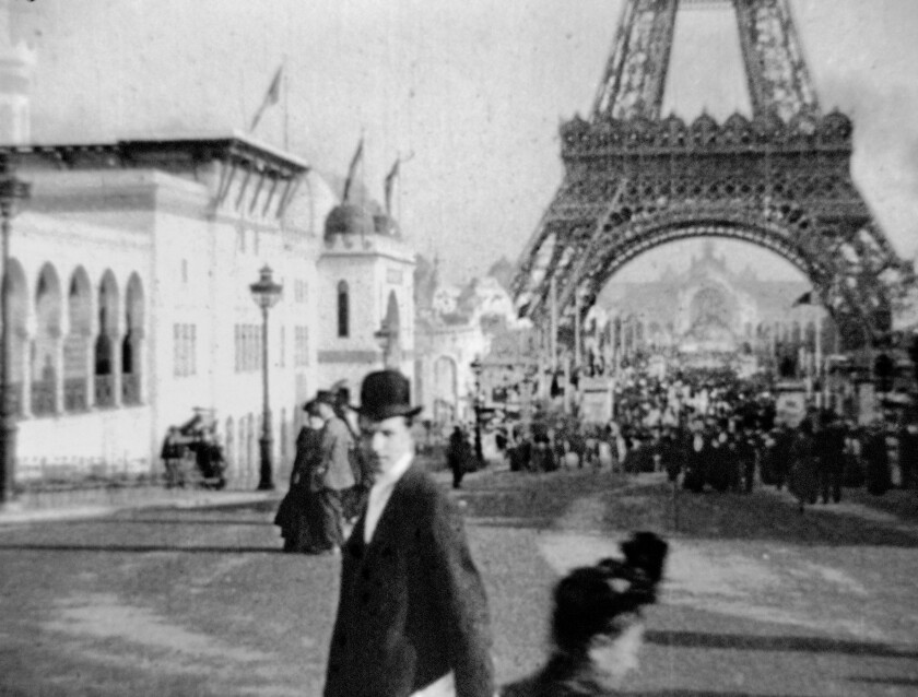 A black-and-white image from 1900 shows a man looking into the camera, with the Eiffel Tower in the background.
