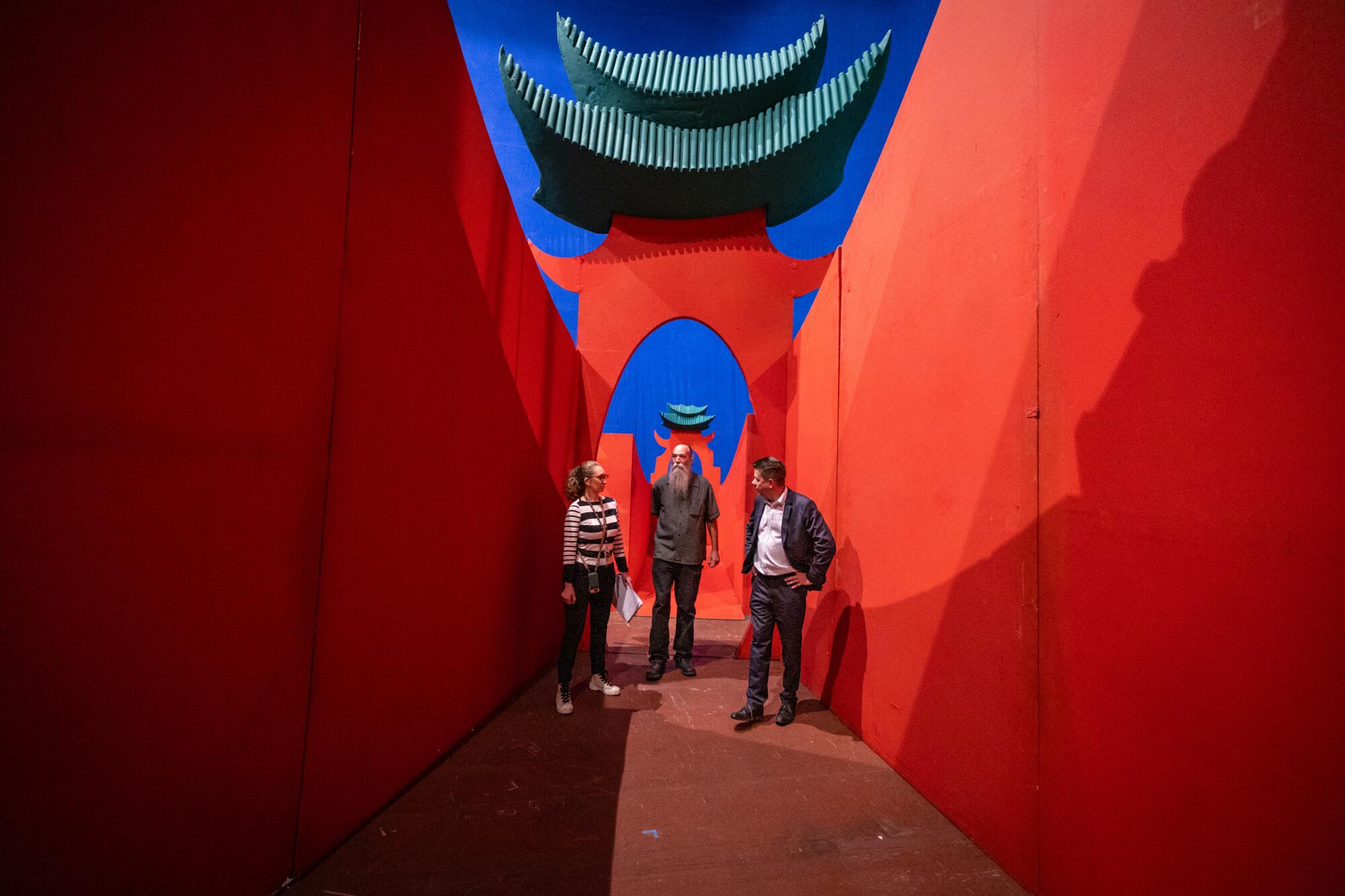 A woman and two men in street clothes talk in front of the mammoth red walls of David Hockney's set for "Turandot."