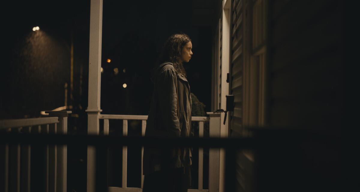 A woman stands on a porch at night as if waiting for someone to answer the door.