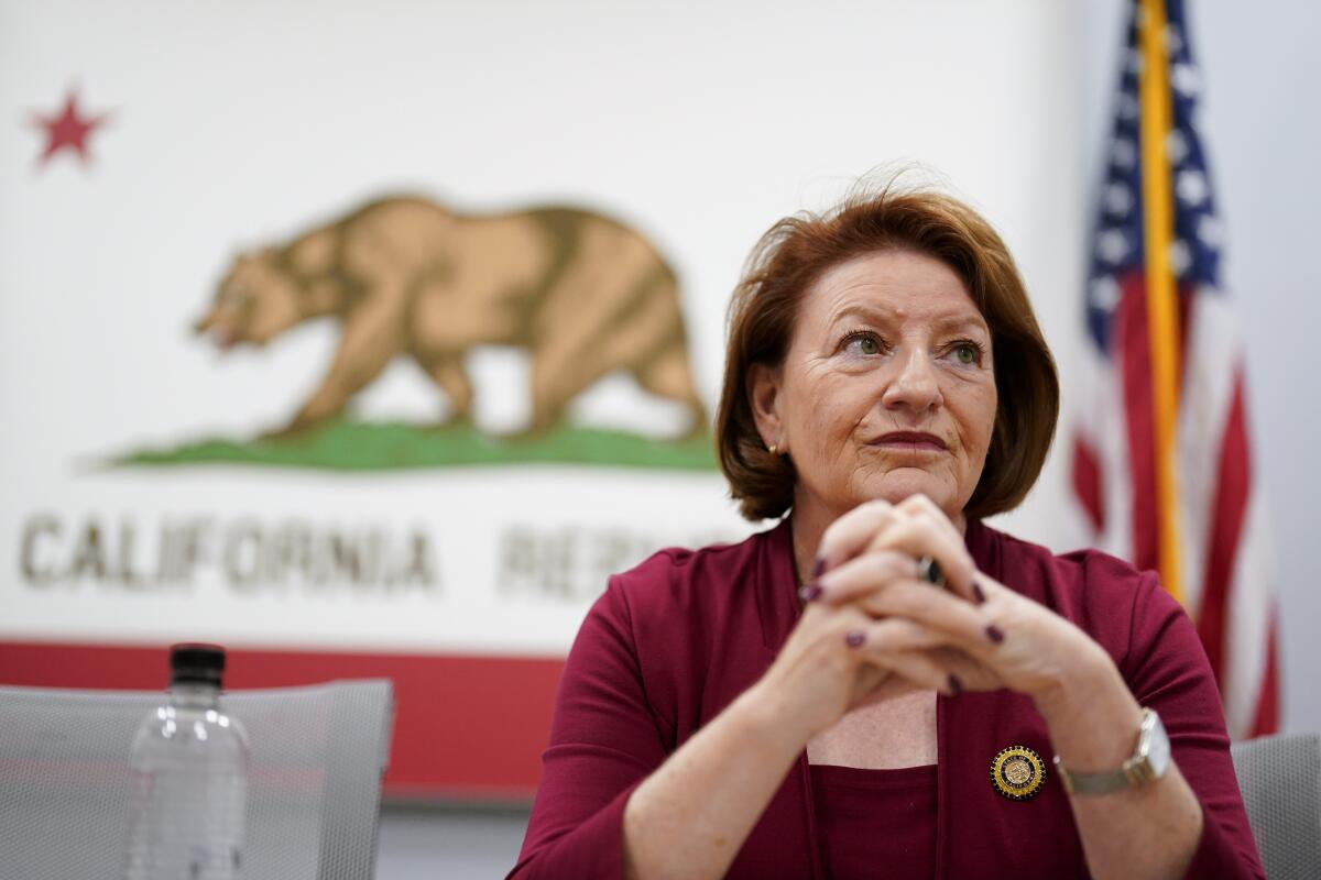 A woman holds her hands together sitting in front of a California state flag.