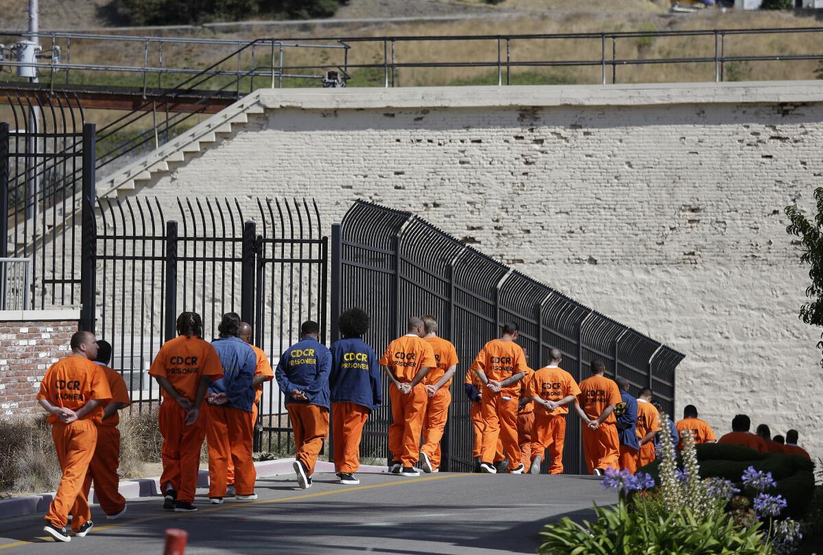 Prisoners in orange jumpsuits with their hands behind their backs walk in a line alongside a fence outdoors