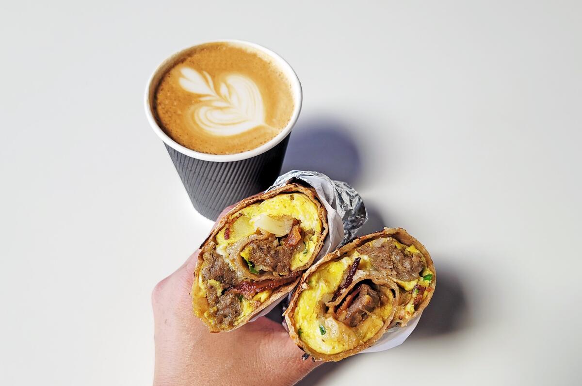 A latte and a hand holding a breakfast chapati, rolled-up flatbread with a filling
