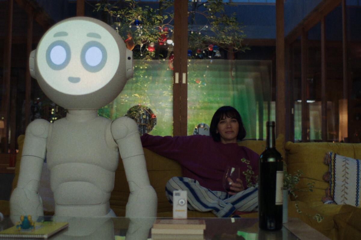 A robot stands near a coffee table as a woman in a red sweater sits on a couch behind it.