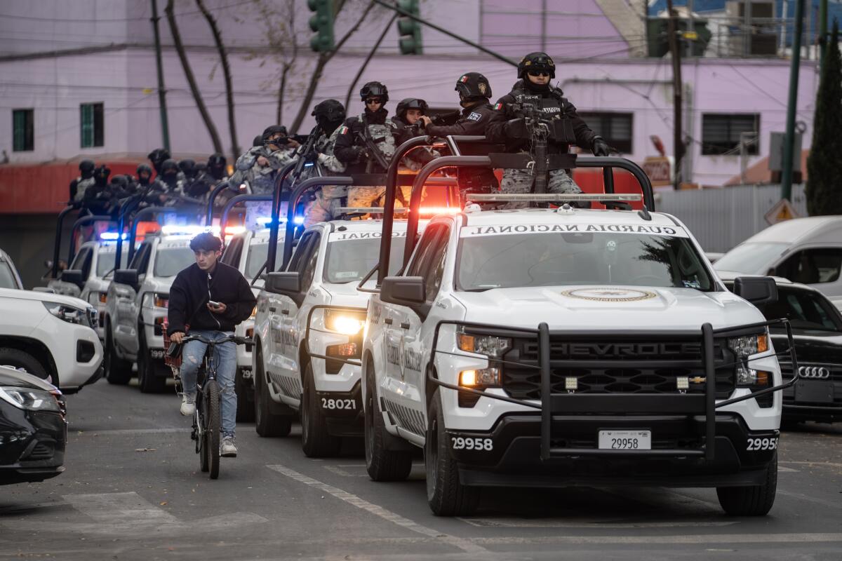 A line of white military trucks, with troops in dark uniforms standing in the truck beds, outside a building