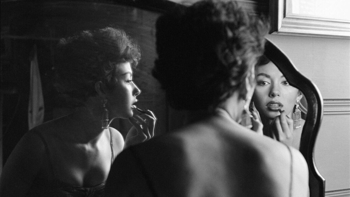 A youthful Rita Moreno is reflected in a mirror as she applies lip pencil.