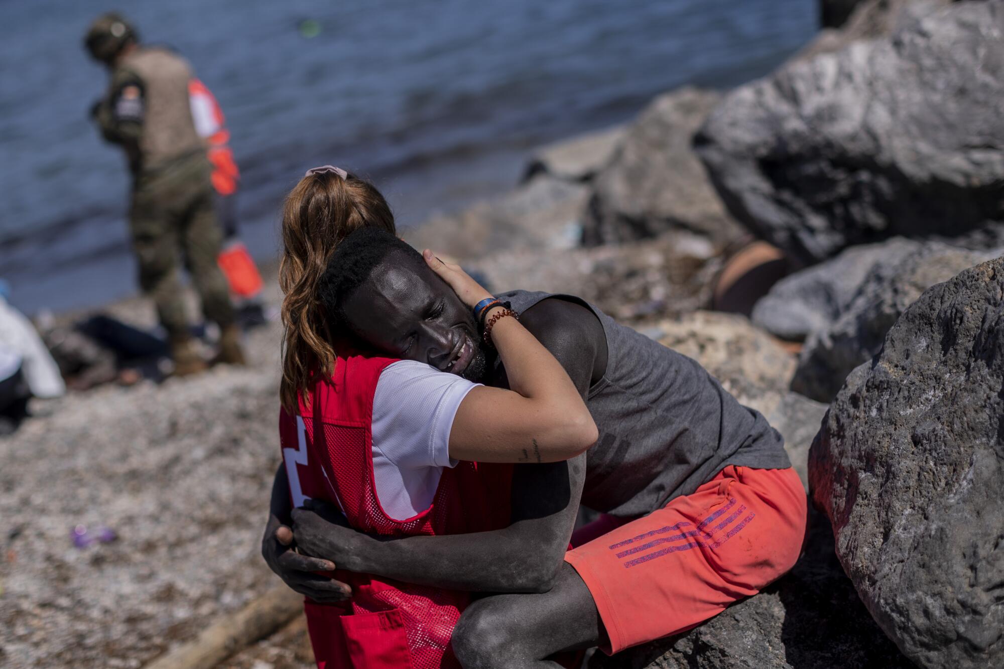 A woman in a Red Cross vest tightly embraces a crying migrant man sitting on a rock at the beach
