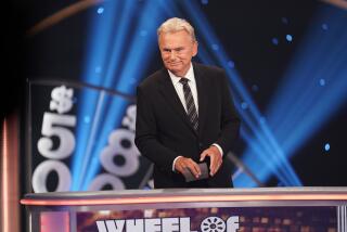Man Pat Sajak standing on Wheel of Fortune stage in black suit, holding cards, smiling and looking off stage