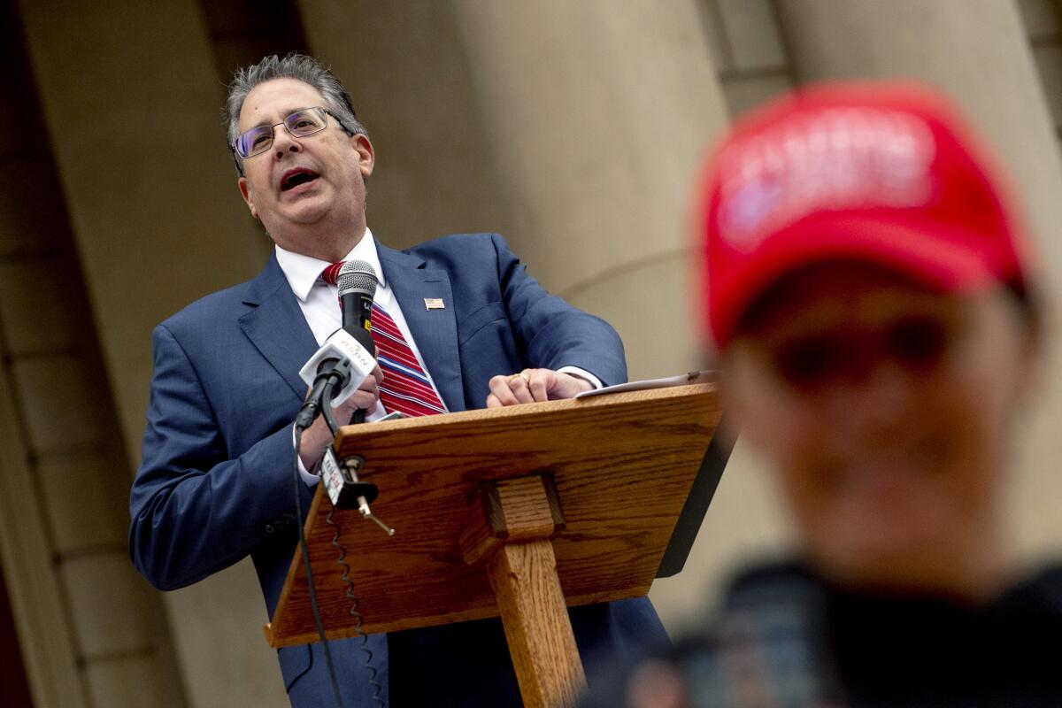 Matthew DePerno speaks at an election rally near a person in a red hat.