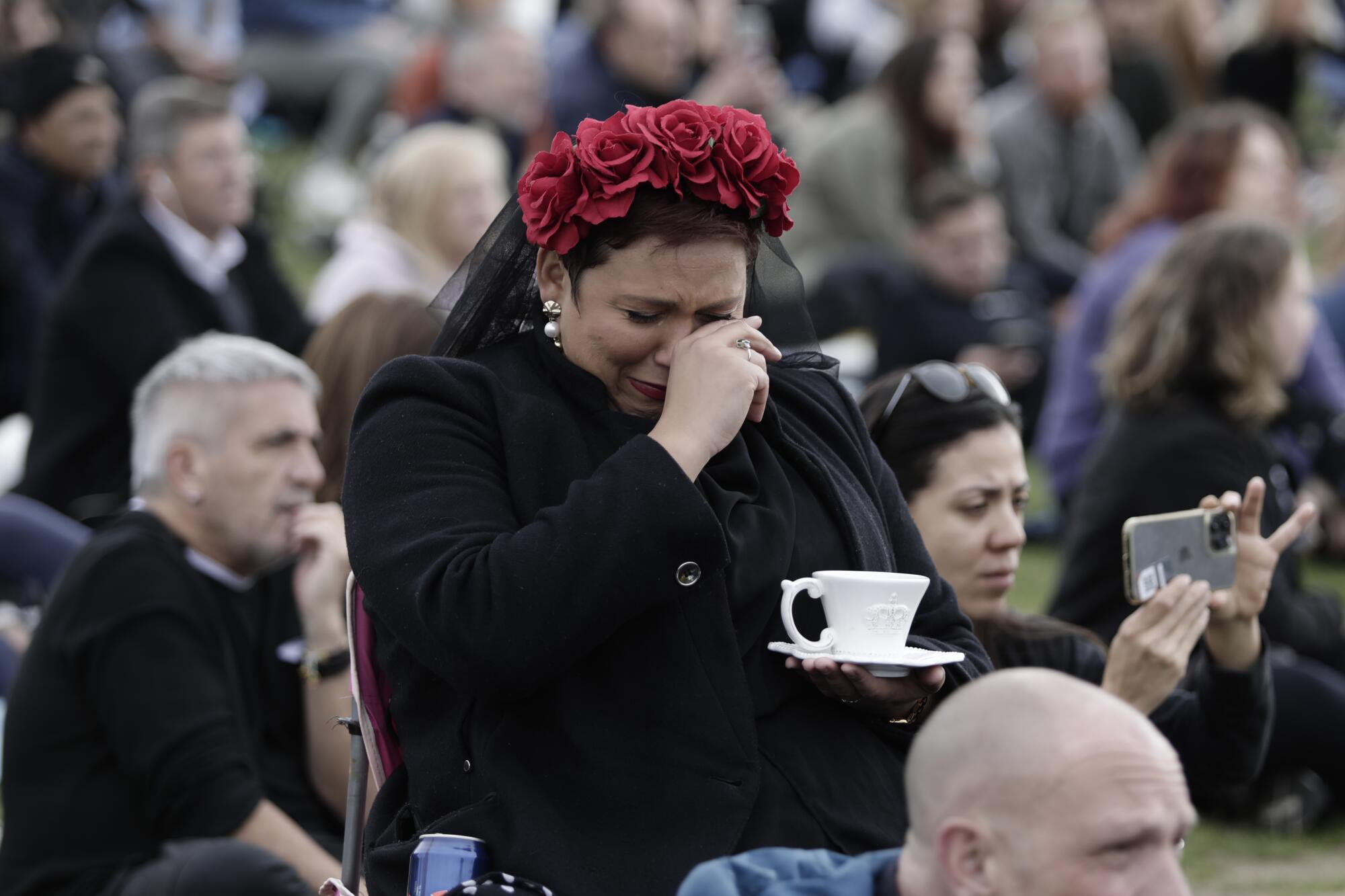A woman in a crowded park holds a teacup and wipes her eyes.