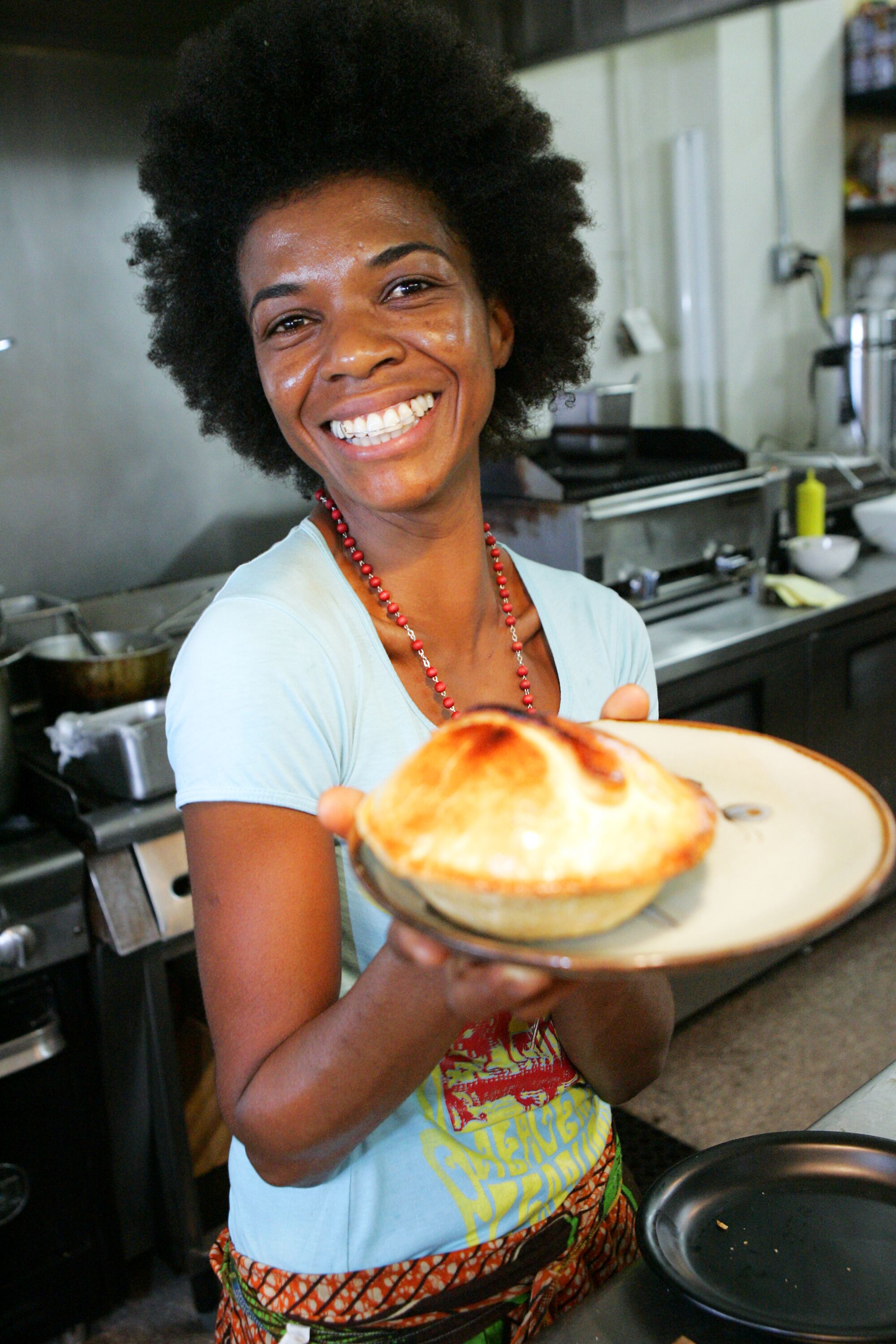 A smiling woman in a restaurant kitchen holds up a chicken pot pie on a plate.