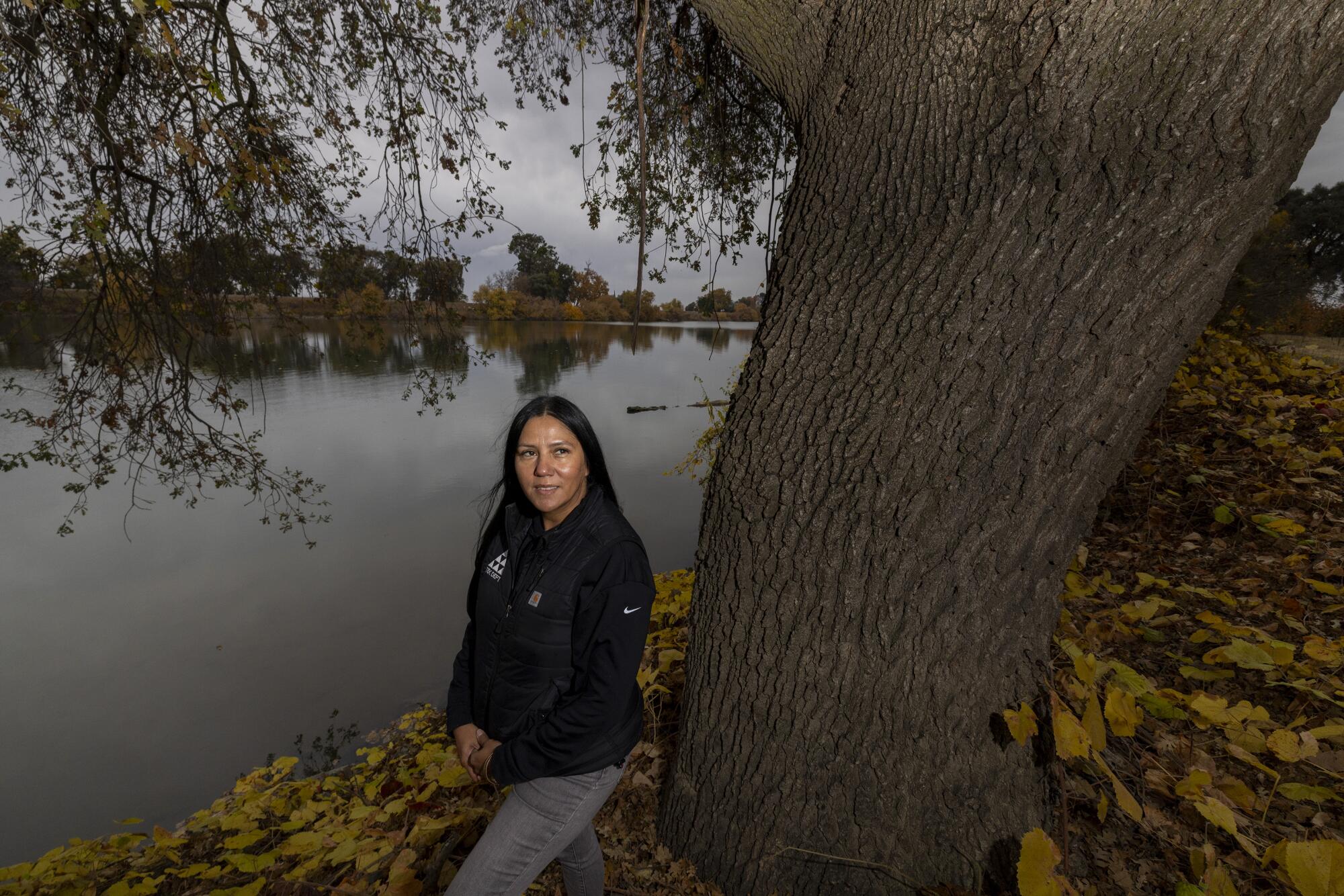 A woman stands next to a tree along a river.
