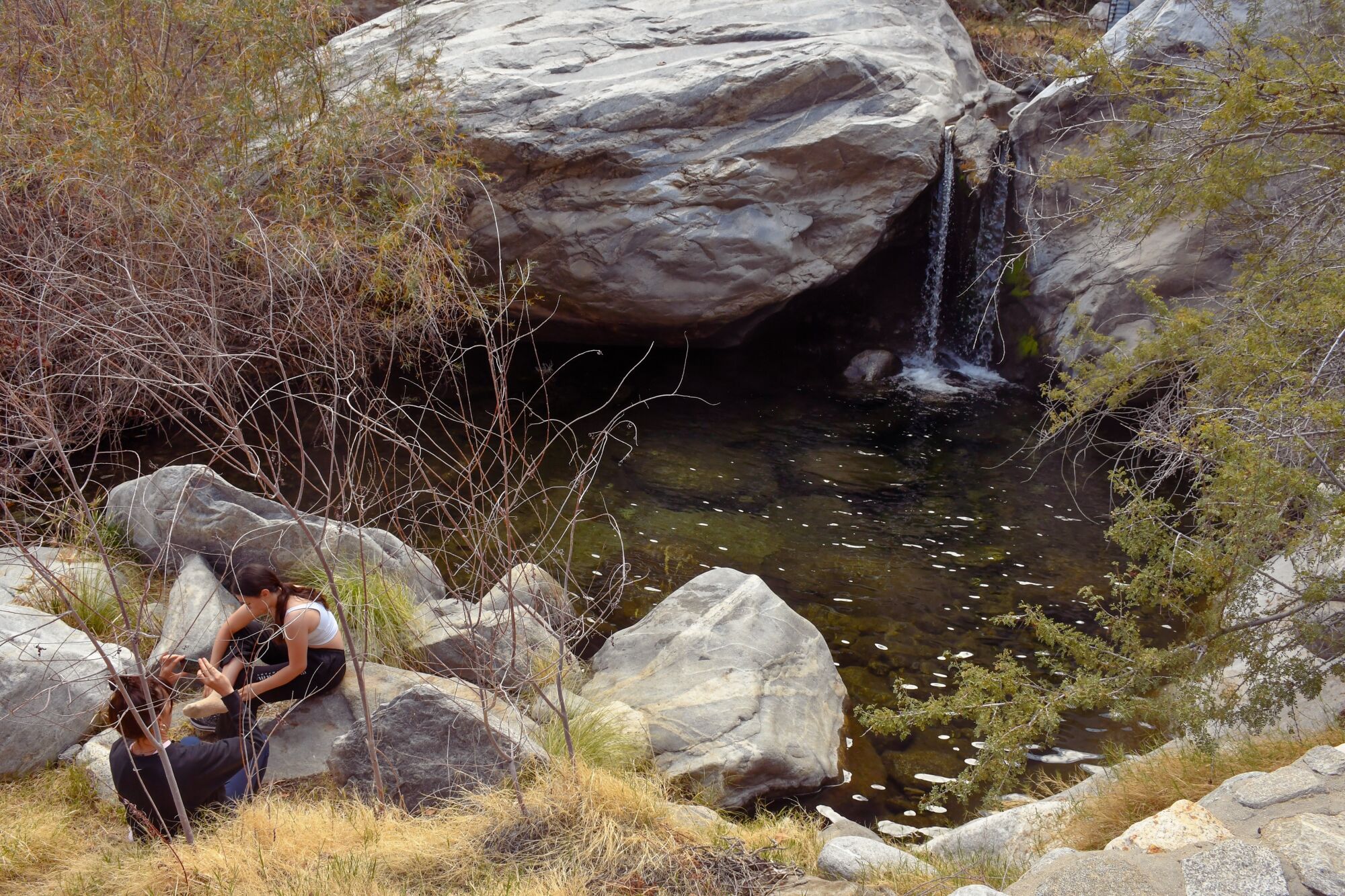 Two people sitting on rocks next to a pool of water and a waterfall