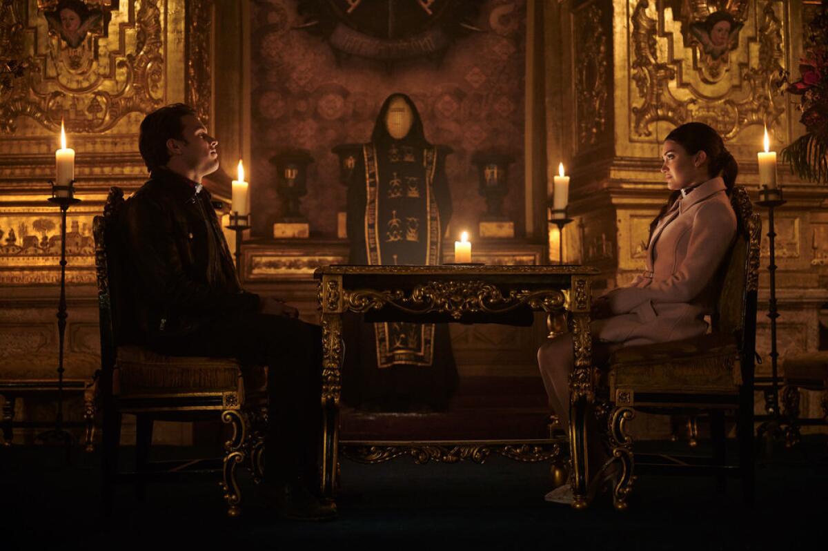 A man and a woman sitting in a gilded, candlelit room across a table from one another