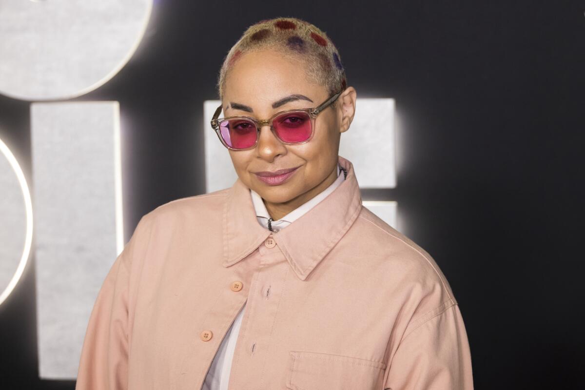 Raven-Symone with a leopard-print buzzcut in a peach pink jacket and red-lensed sunglasses smiling at a red carpet