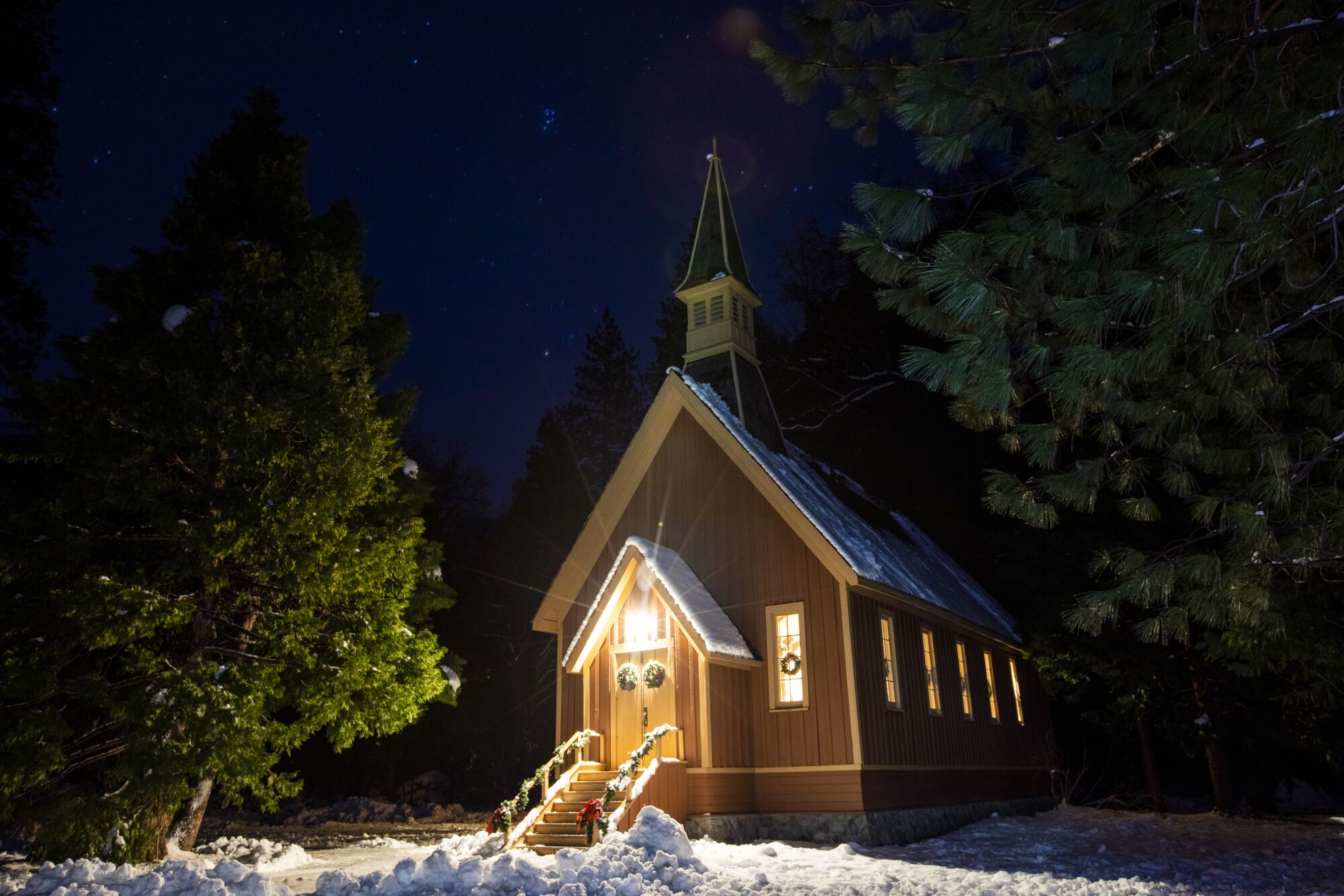 Light shines from a small, many-windowed building with a steeple flanked by evergreens under a night sky.