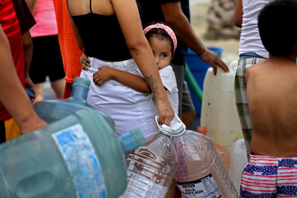 A young girl clings to a person holding empty plastic jugs as they wait in line for water 