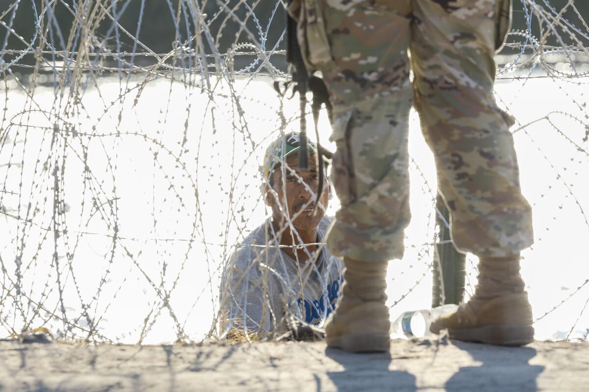 A man stands behind razor wire looking up at a person seen from the legs down in a camouflage uniform and boots.