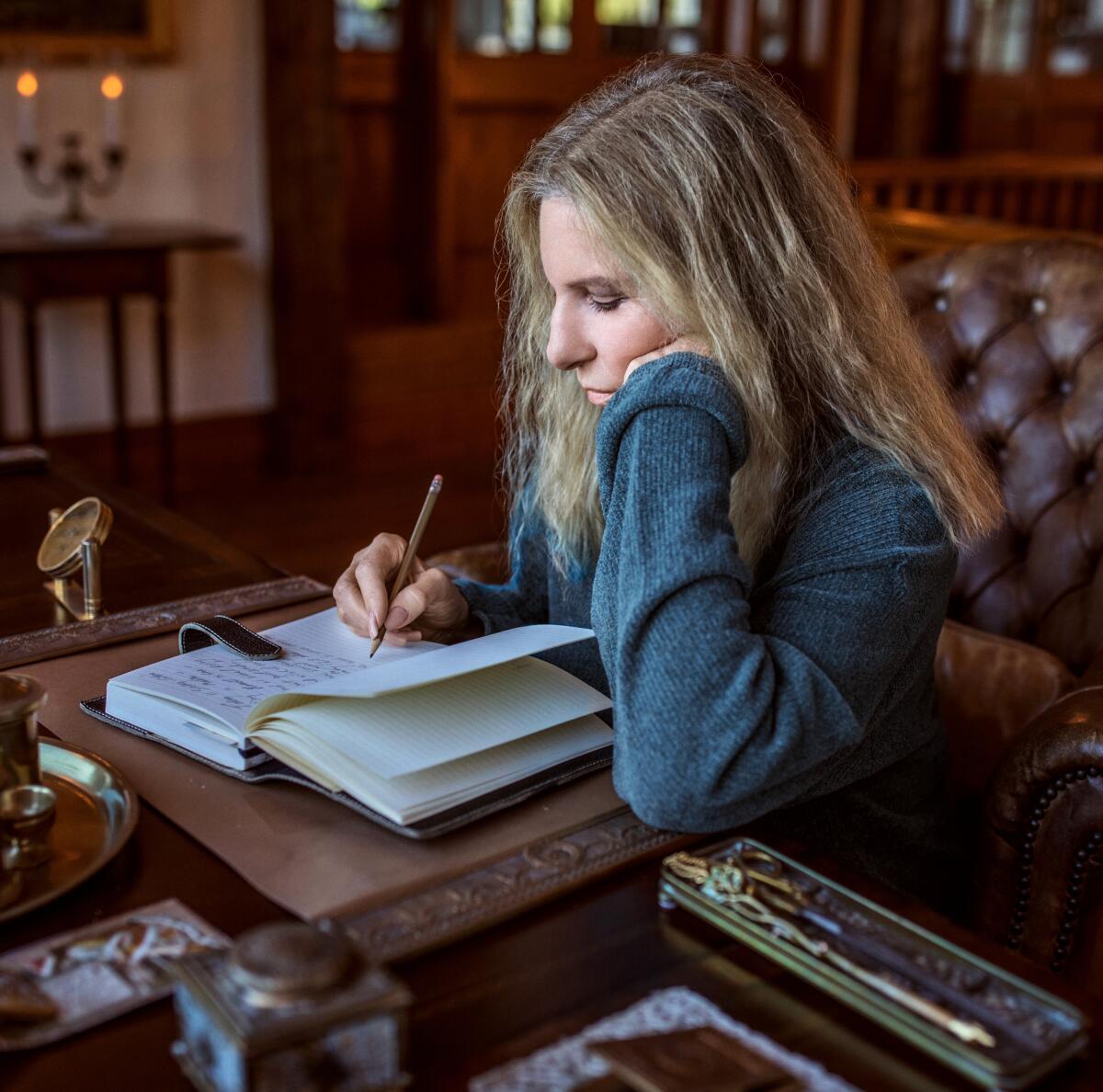Barbra Streisand sits at a desk writing in a notebook.