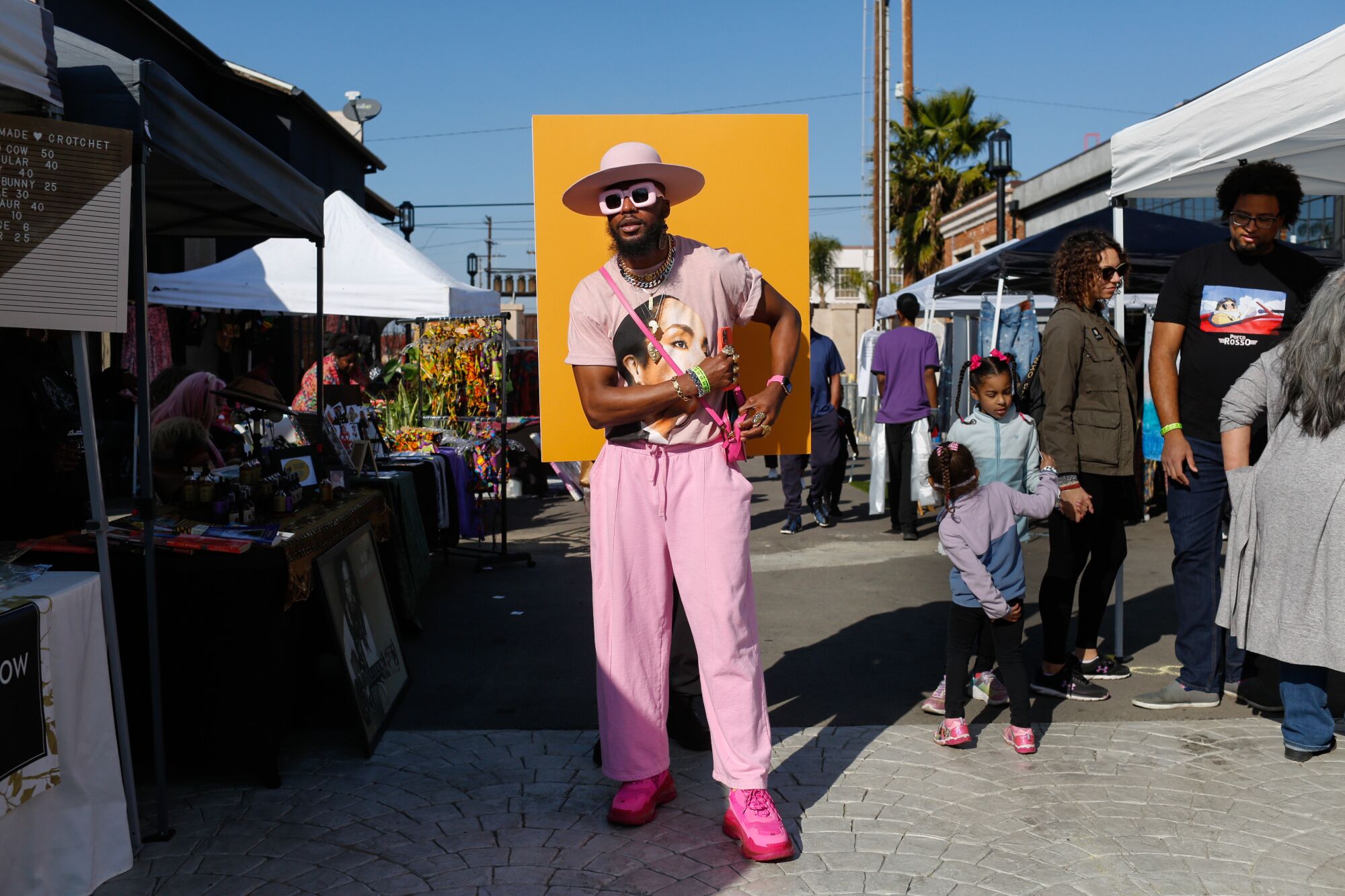 A person in pink pants and shoes, printed T-shirt and broad-brimmed white hat stands at an outdoor flea market.