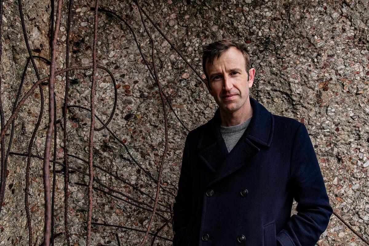 A man, standing in nature and wearing a blue coat, looks at the camera.