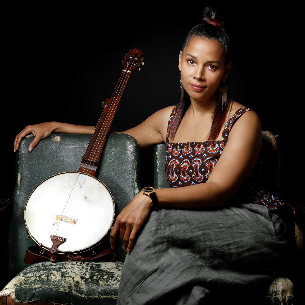 Rhiannon Giddens, wearing a brightly patterned top and grey pants, sits next to a banjo.