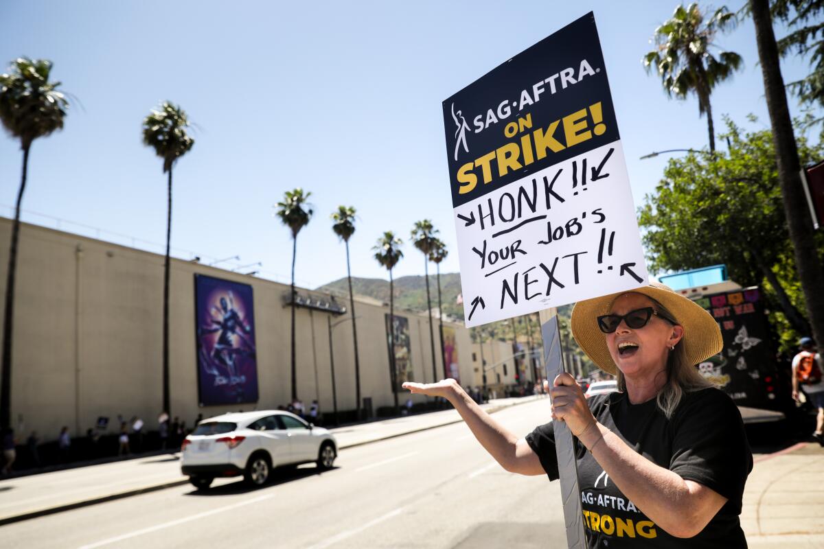 A woman in a straw hat holds a SAG-AFTRA picket sign outside the Warner Bros. studio lot.
