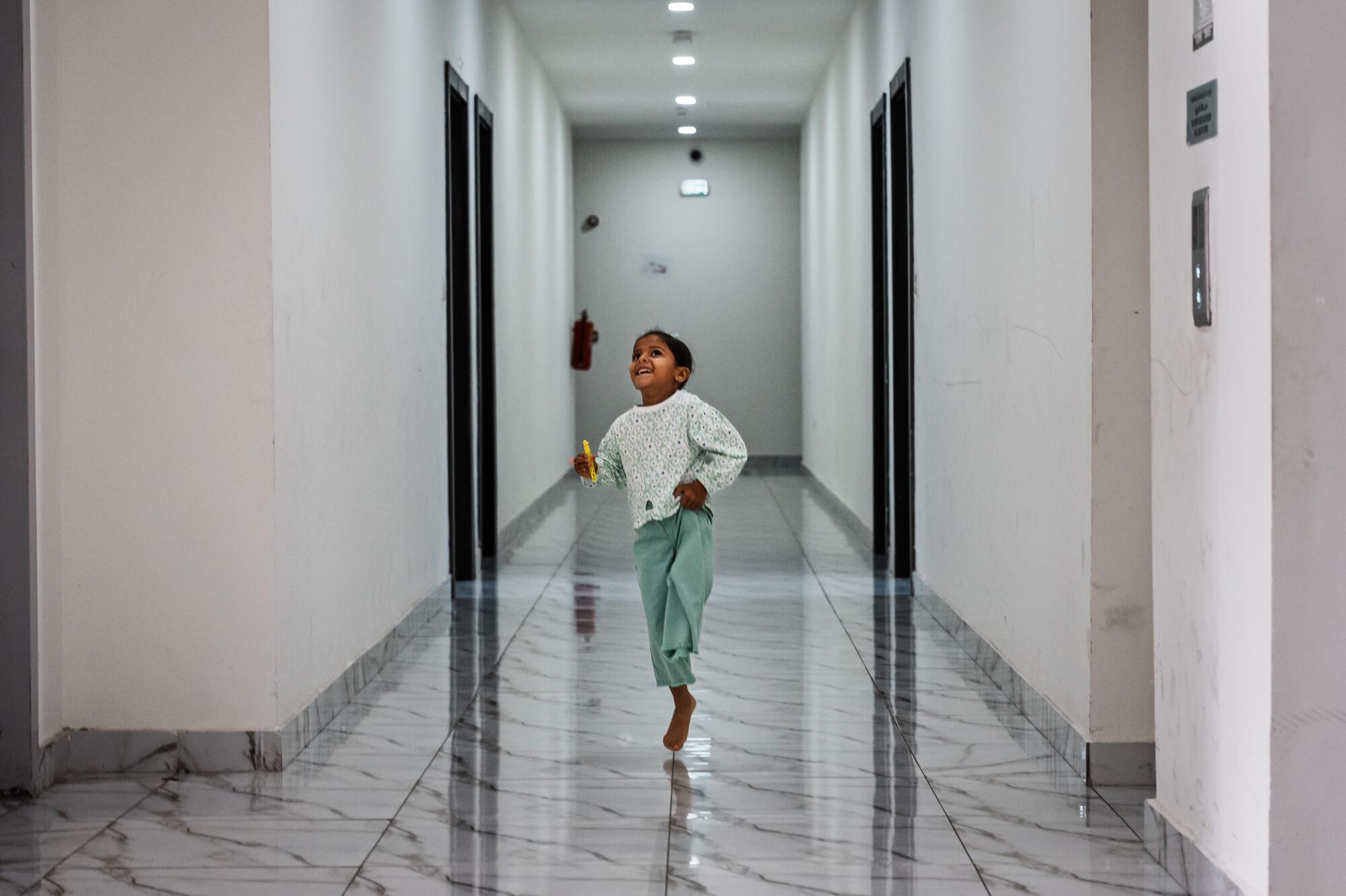 A child in a light green top and green pants hops barefoot on one leg in a hallway 