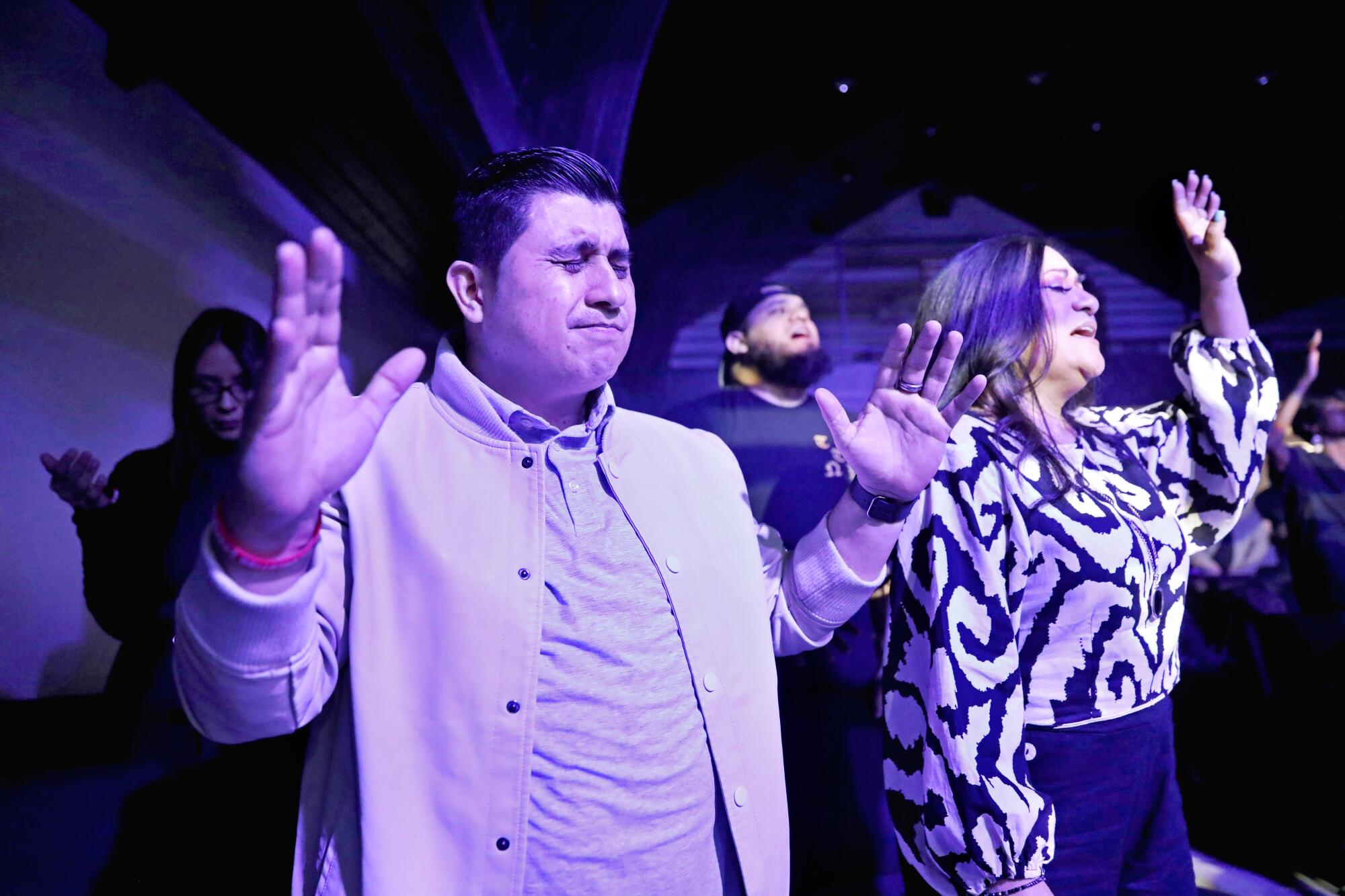 A few people standing, bathed in purple light, with their eyes closed and hands raised