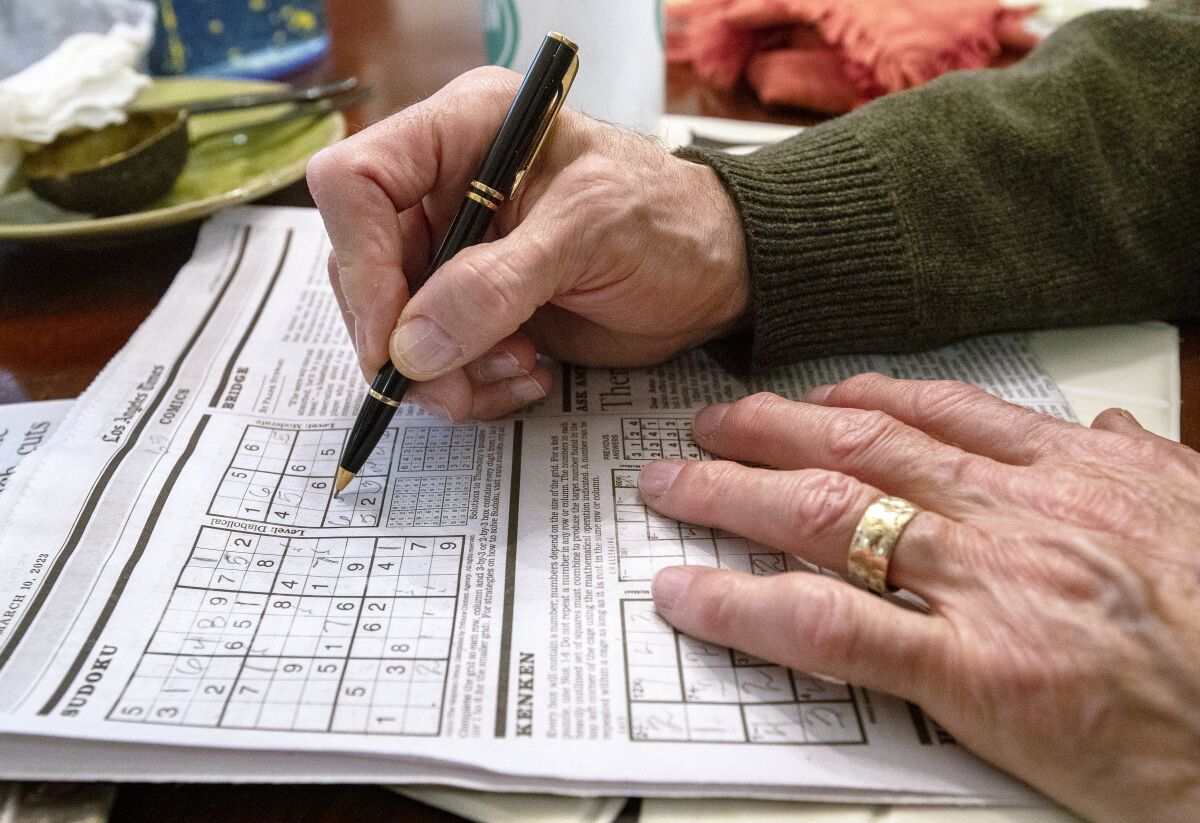 Les mains reposent sur une page de journal qui affiche des mots croisés et des puzzles Sudoku. 