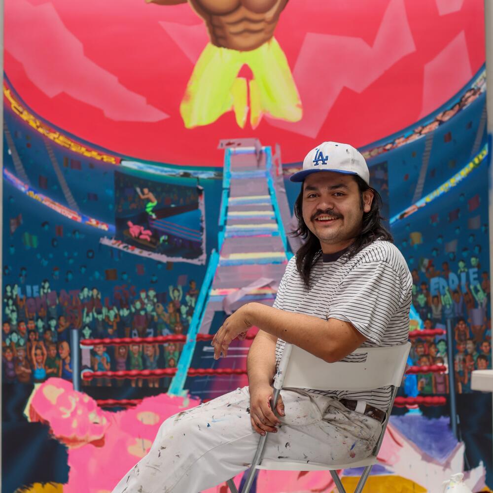 A smiling long-haired man in a baseball cap sits in front of artwork.