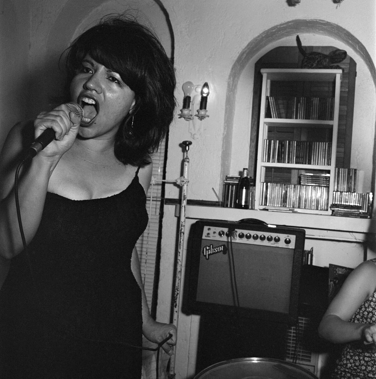 A woman in a black dress sings into a microphone before an amplifier set up in an apartment.
