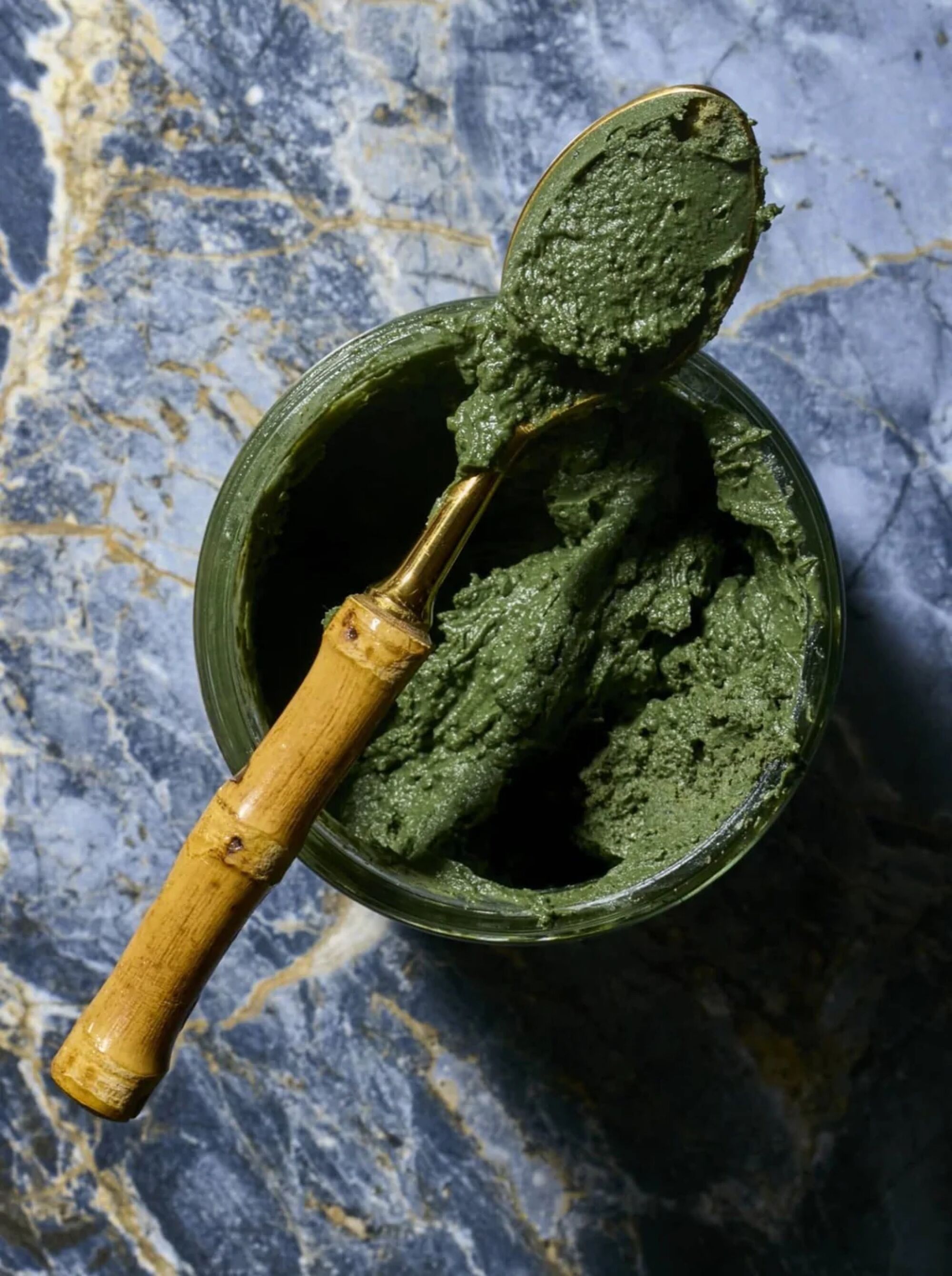 a top-down view of a bamboo spoon lying across a jar filled with green mud clay that sits on top of a marble surface