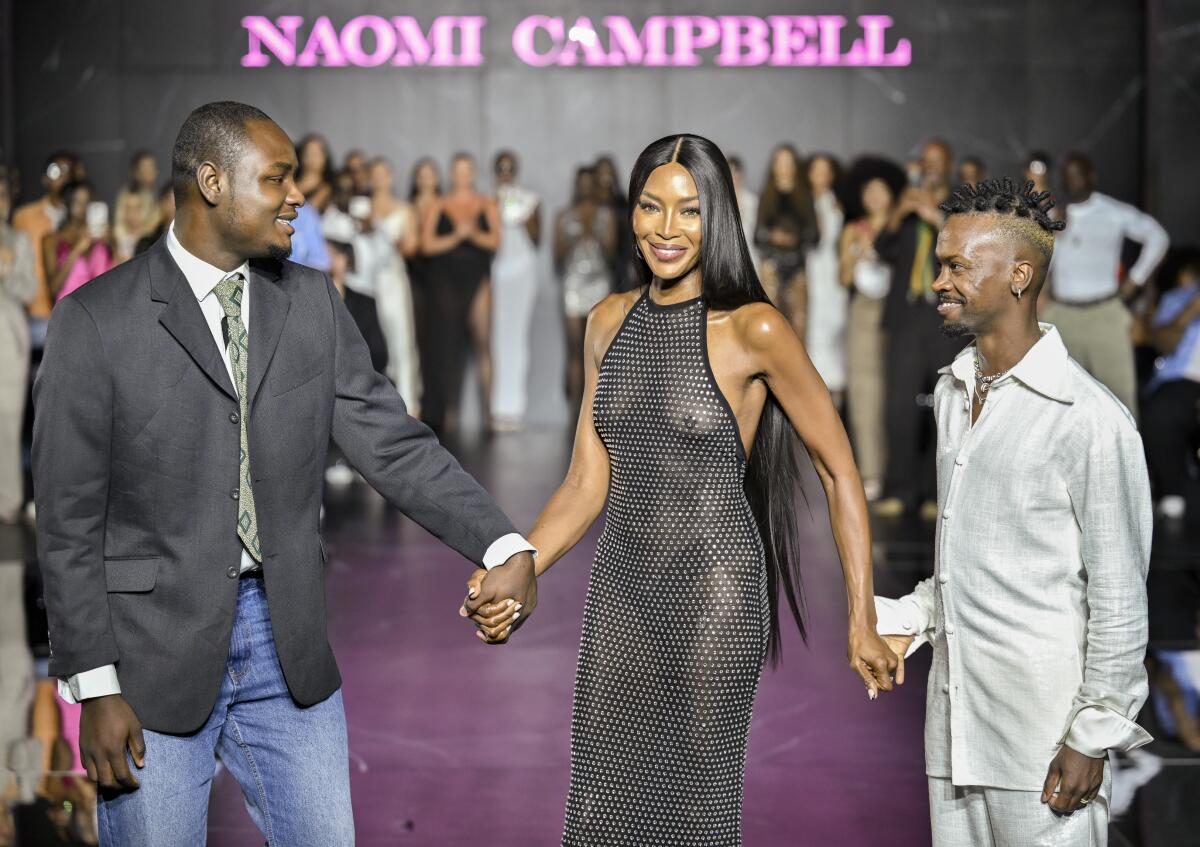 Naomi Campbell smiles while wearing a sheer black dress and holding hands with Victor Anate and Edvin Thompson on a runway.