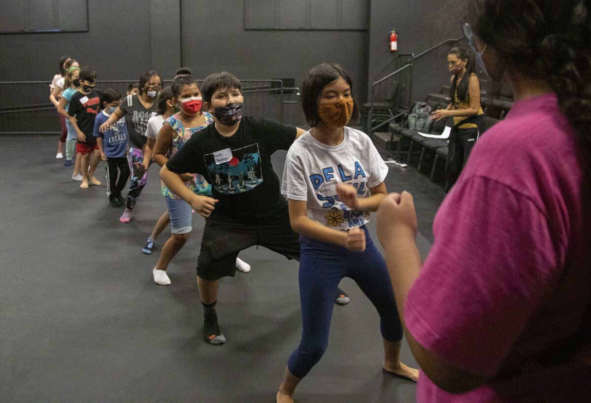 Students wearing masks are seen rehearsing choreography inside a black box theater.
