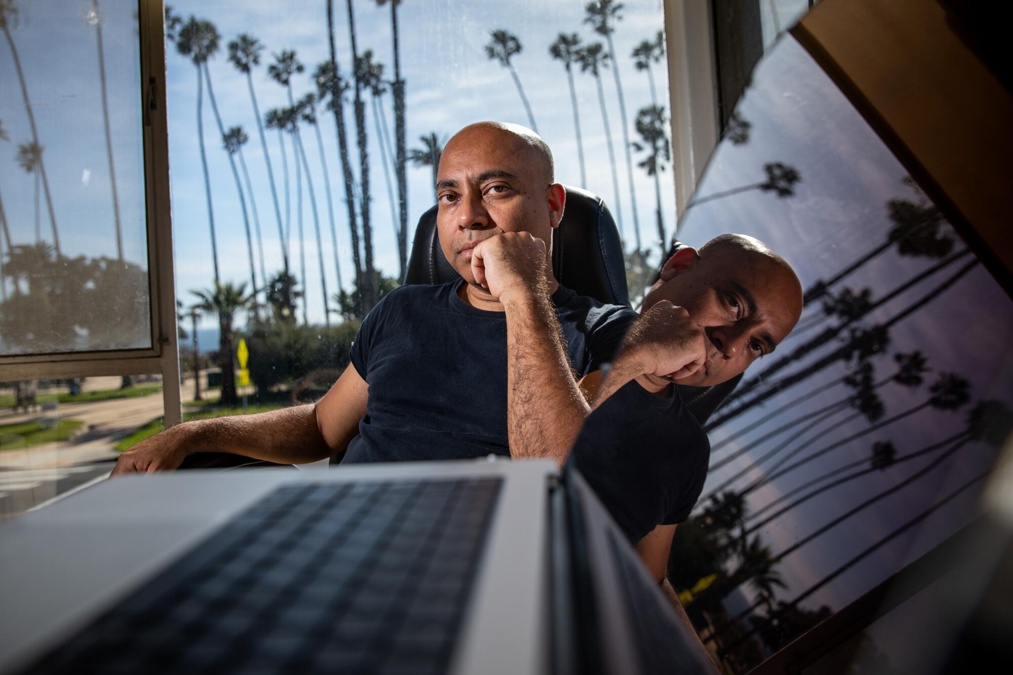 A man sits with a laptop open in front of him and palm trees visible behind him.