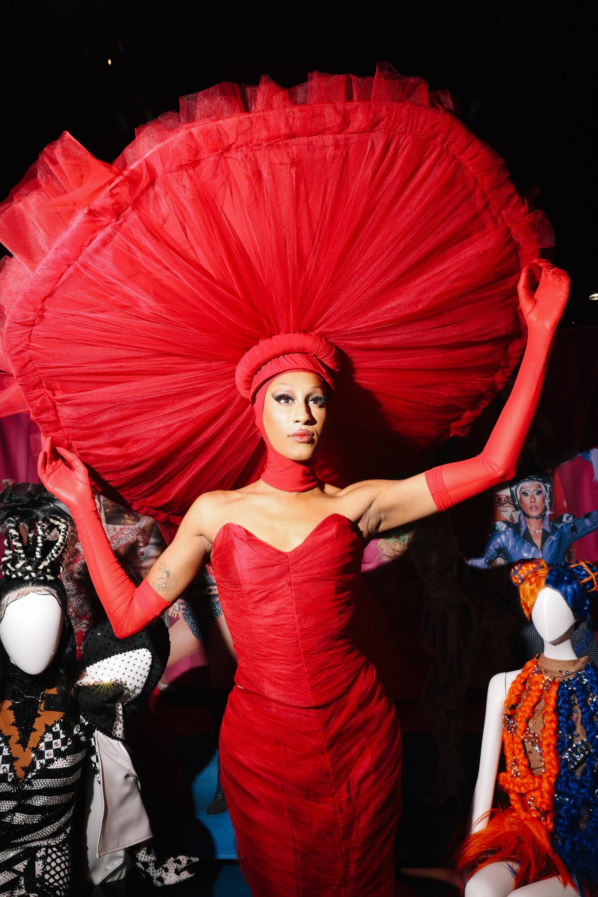 A person in drag poses with their hands on their huge hat.