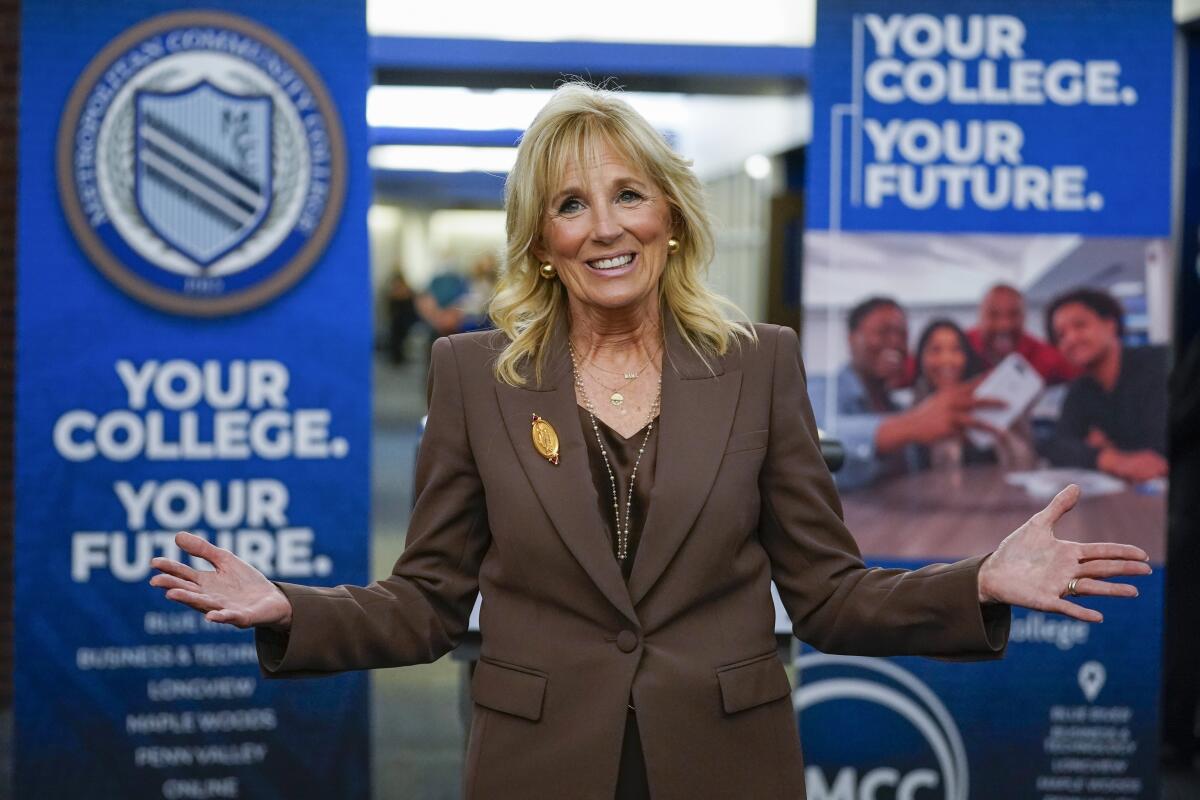 Jill Biden, in a brown suit, stands in front of signs that read "Your college. Your future."