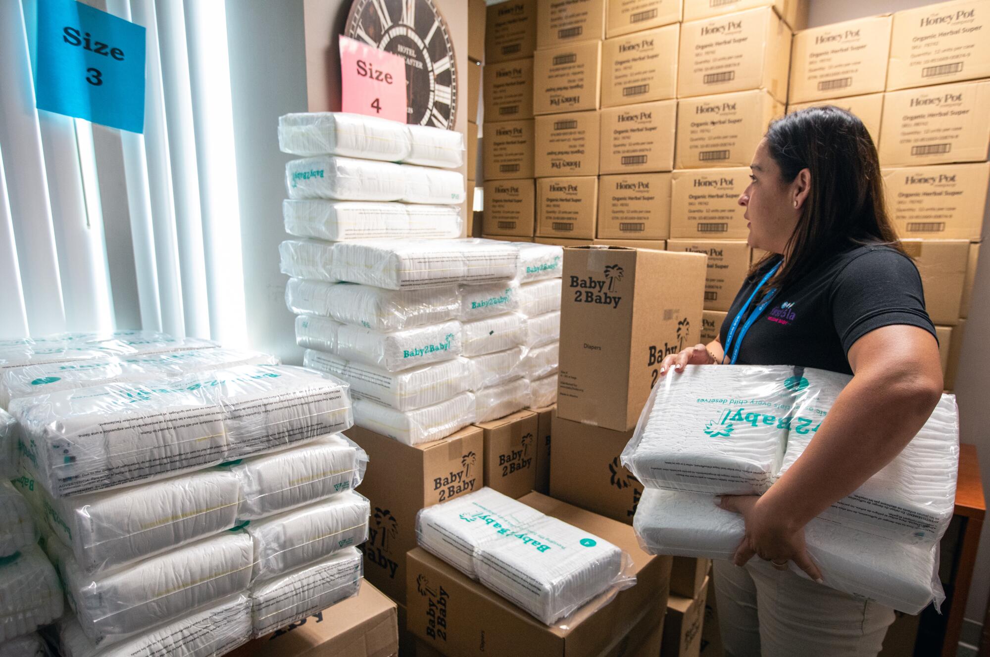 A woman holding white packages looks at stacks of similar packages next to stacked boxes 