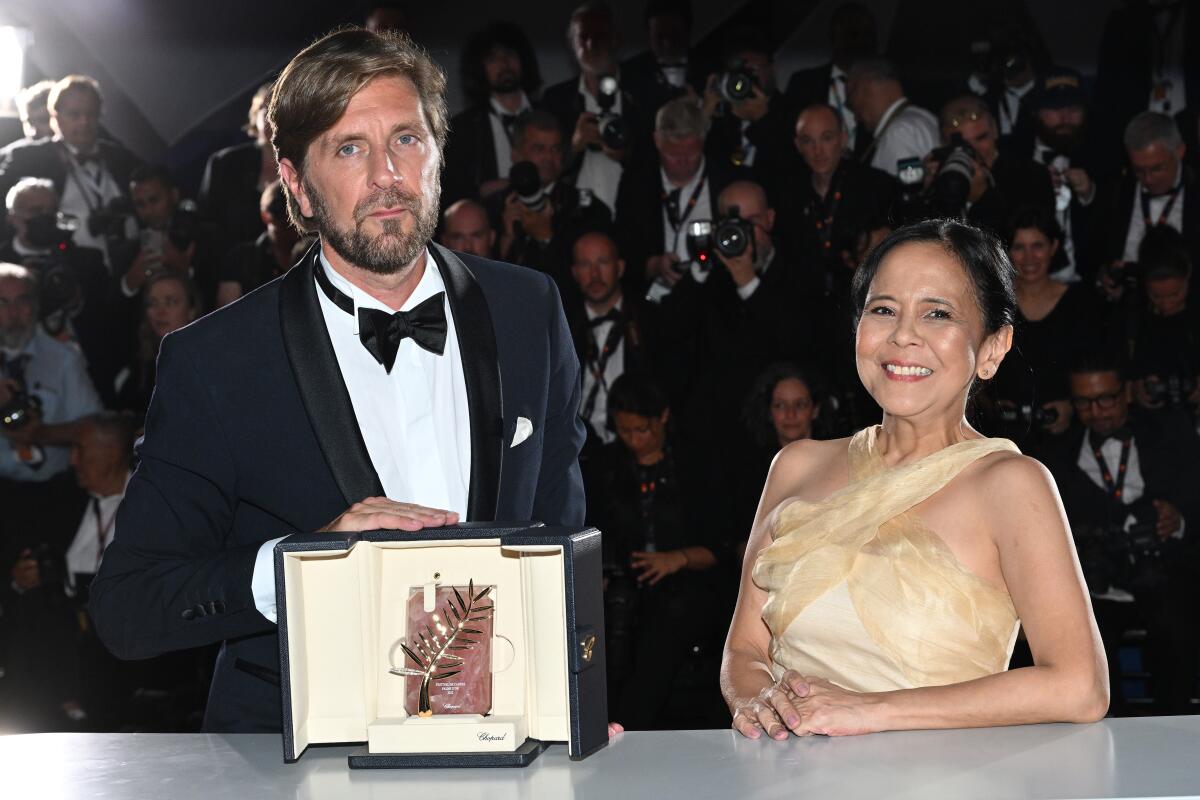 A man in a tuxedo and a woman in a formal dress pose with an award before a crowd of photographers.