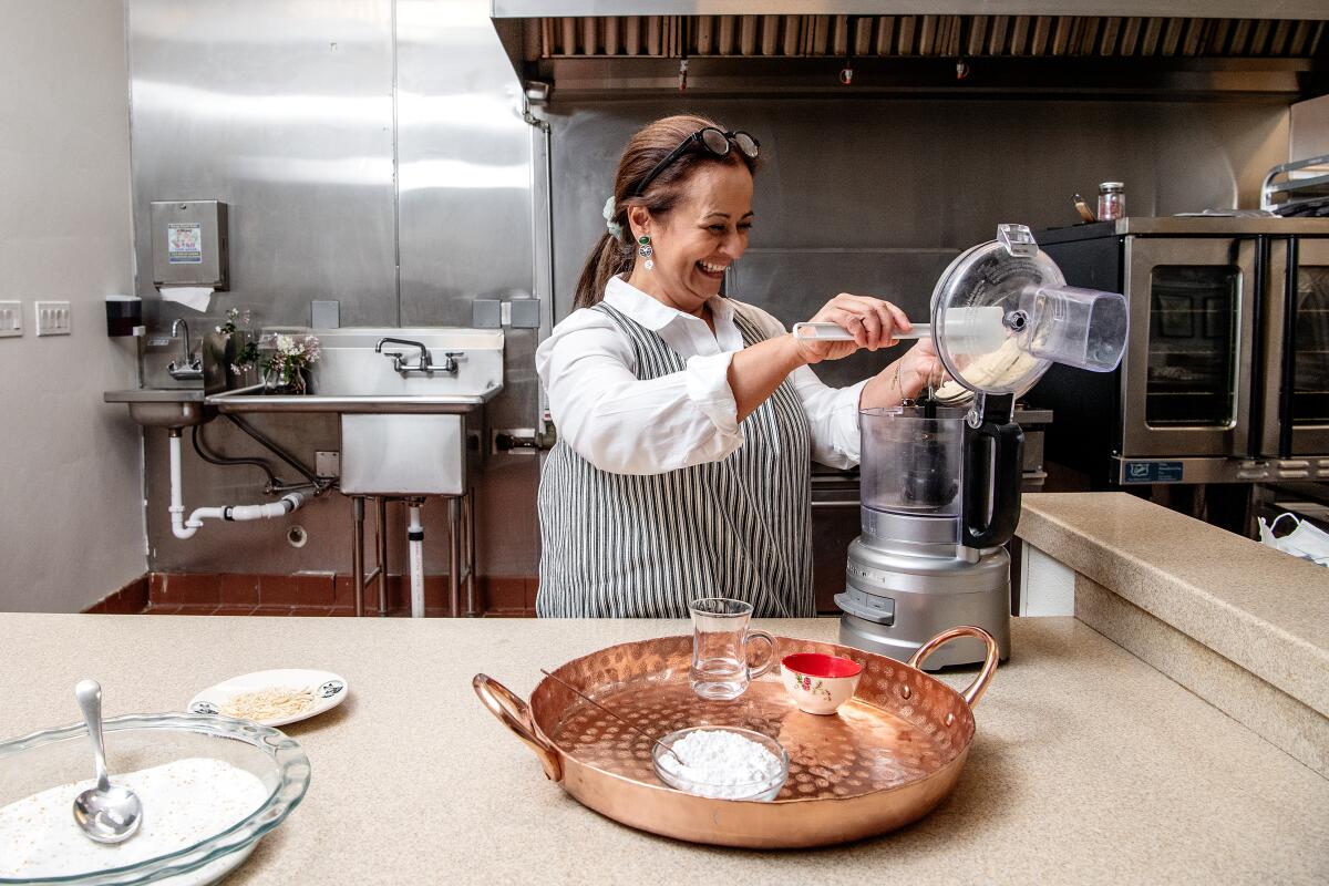 A smiling woman in an apron pours ingredients into a blender in an industrial kitchen.